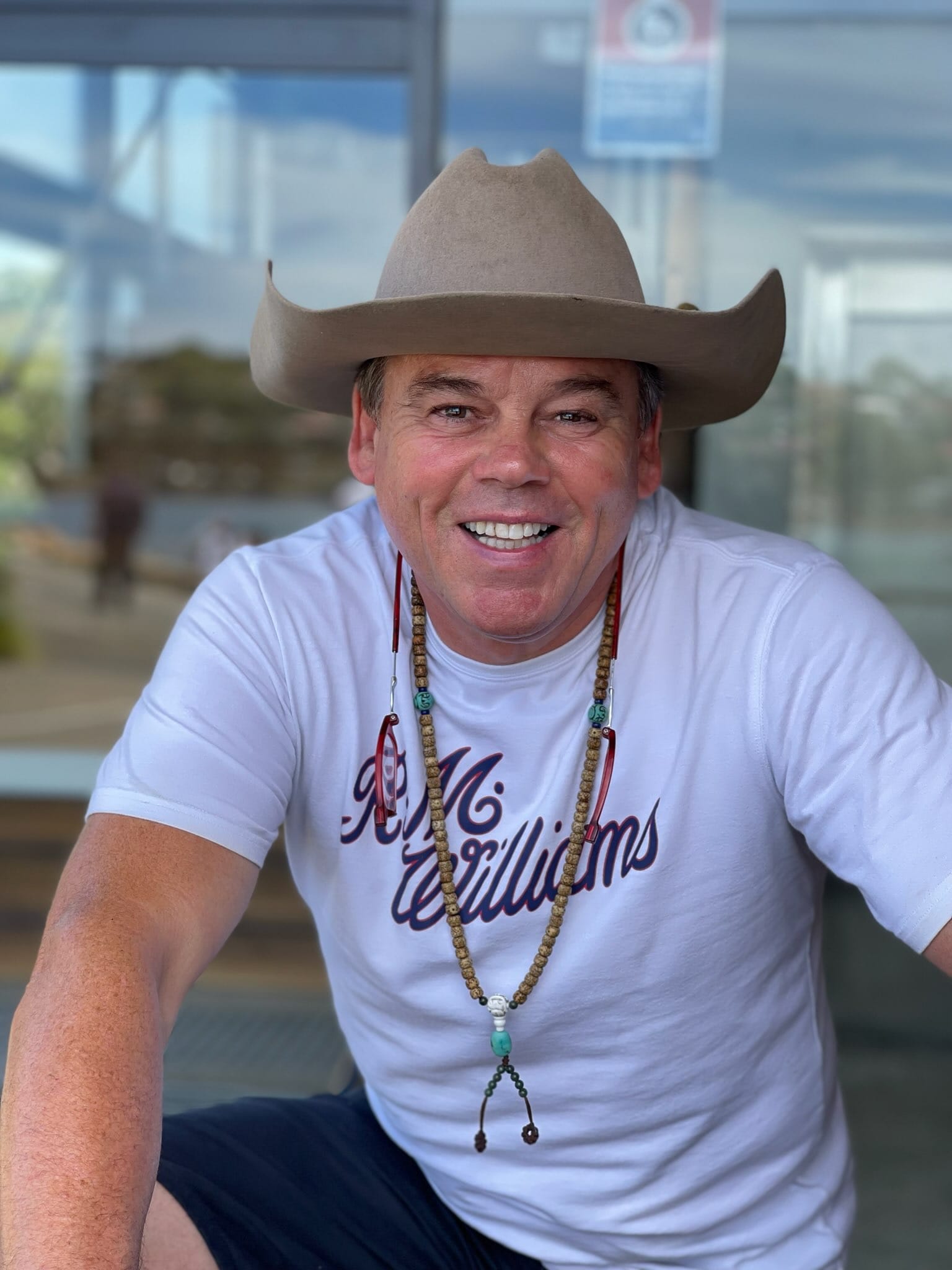 Smiling man wearing a beige cowboy hat, white T-shirt, and beaded necklace outdoors.