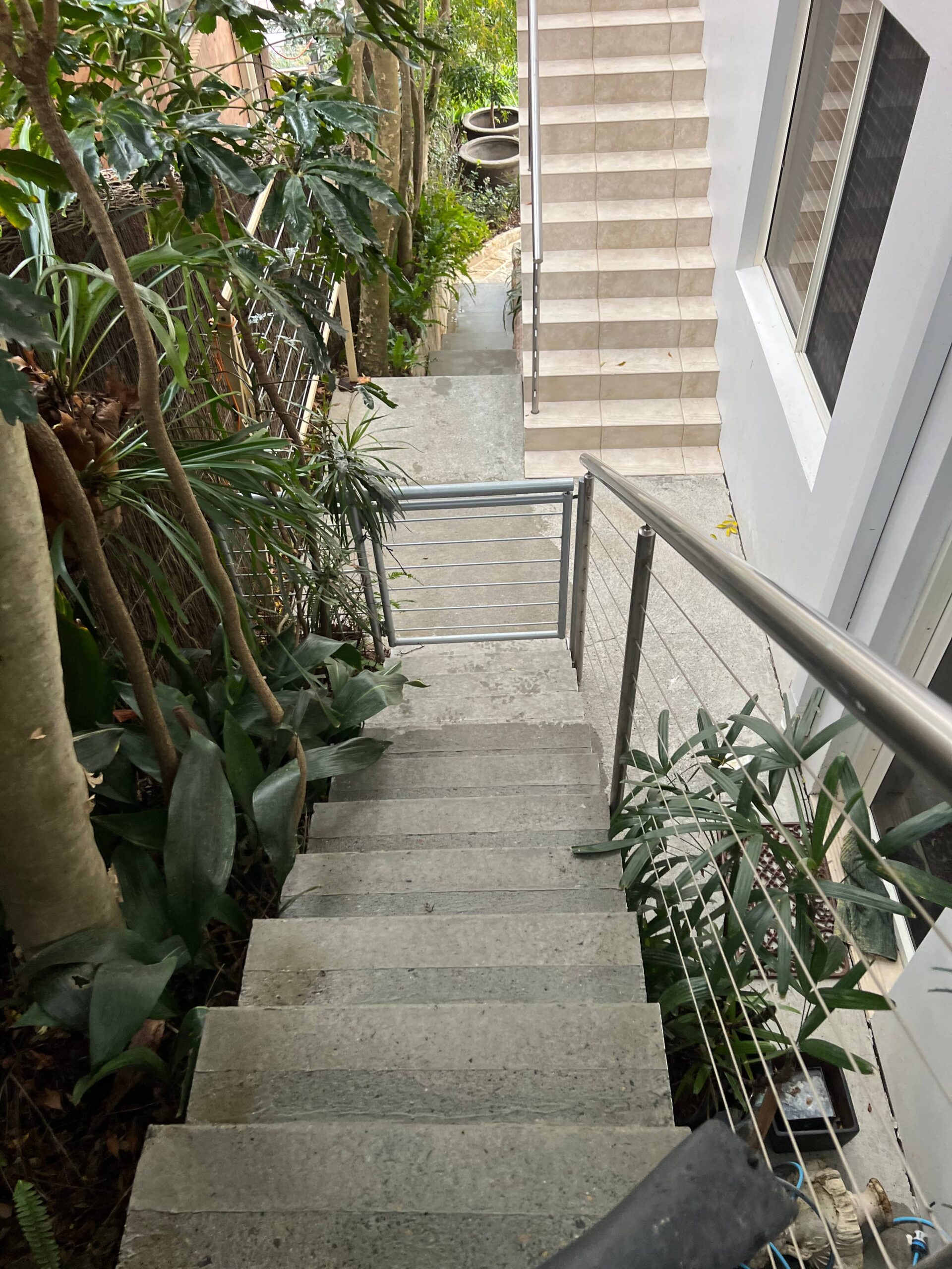Concrete outdoor staircase with metal railings and plants, leading down to a walkway near a white building.