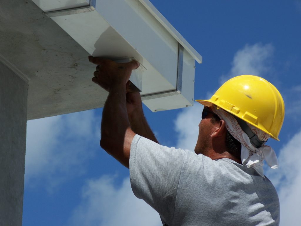 Construction worker in yellow hard hat installs or repairs white soffit, ensuring gutter clearance, clear blue sky.