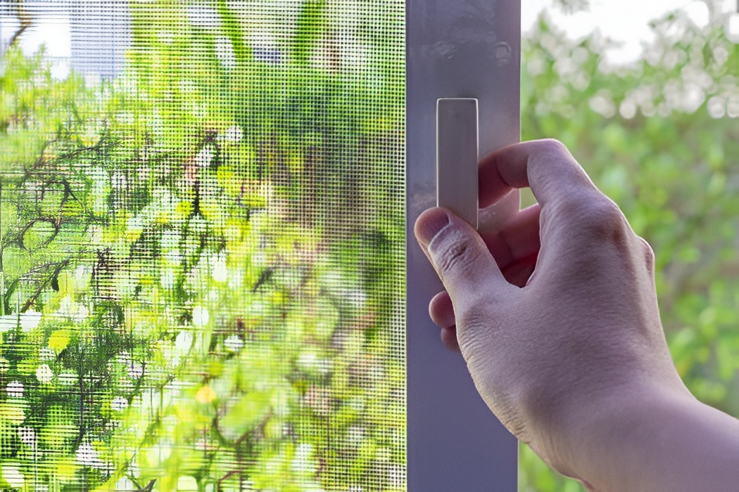 Hand opens window with mesh, hinting at fly screen repair need, with green foliage visible outside in natural daylight.