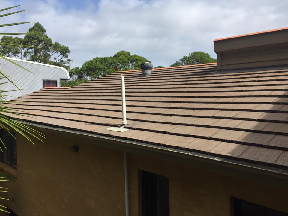Brown tiled roof with two vents, a white pipe, and clean gutters—great for gutter cleaning—trees and sky in background.