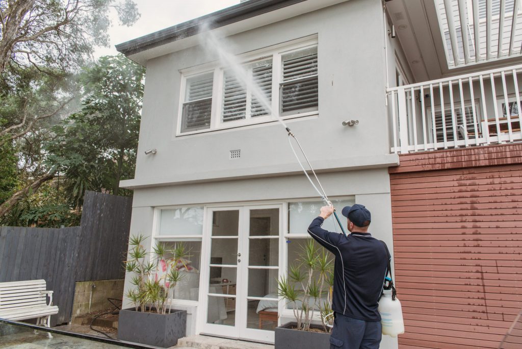 Person washing a two-story house exterior with pressure washer, cleaning white walls and glass doors efficiently.