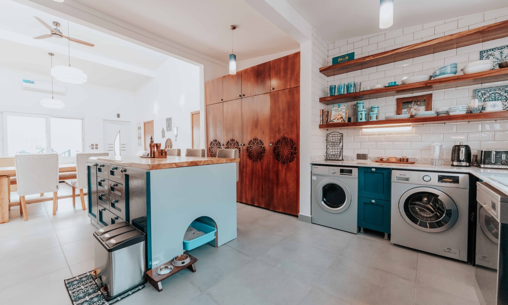 Bright open-plan kitchen with a blue island, wooden cabinets, and white subway-tile walls; laundry appliances (washing machine and dryer) beneath a marble countertop to the right; open wooden shelves with dishes above the counter.