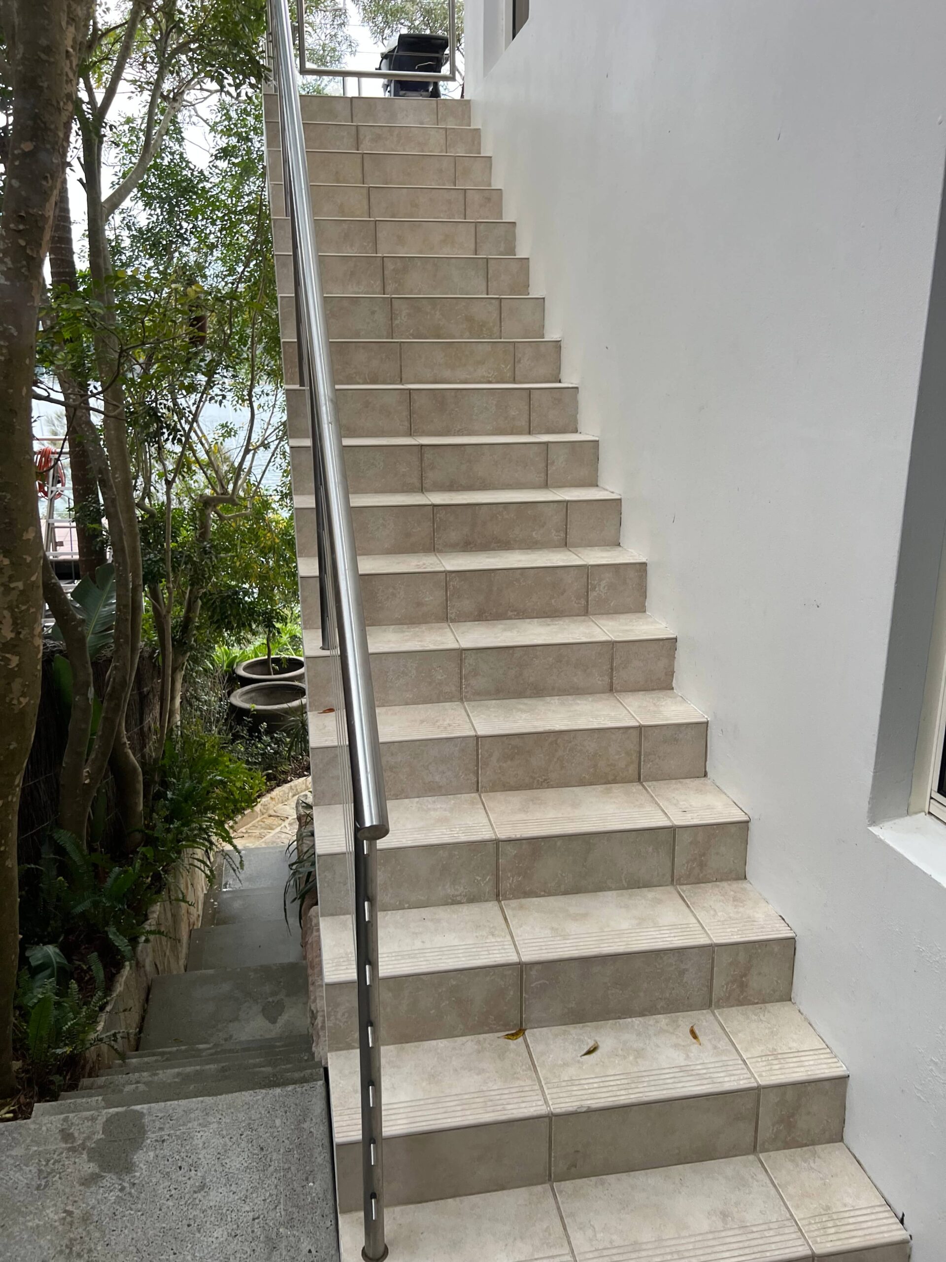 Outdoor tiled staircase with metal handrail beside a white wall, surrounded by trees and plants.