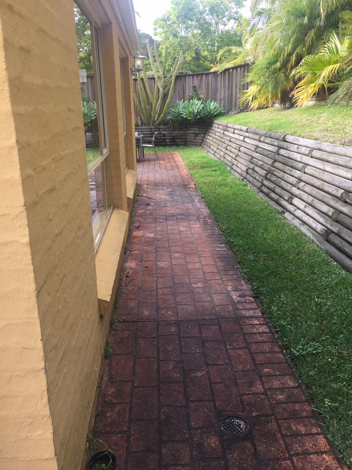 Pressure Cleaning Service for brick pathway along yellow house bordered by grass, wall, and lush plants.