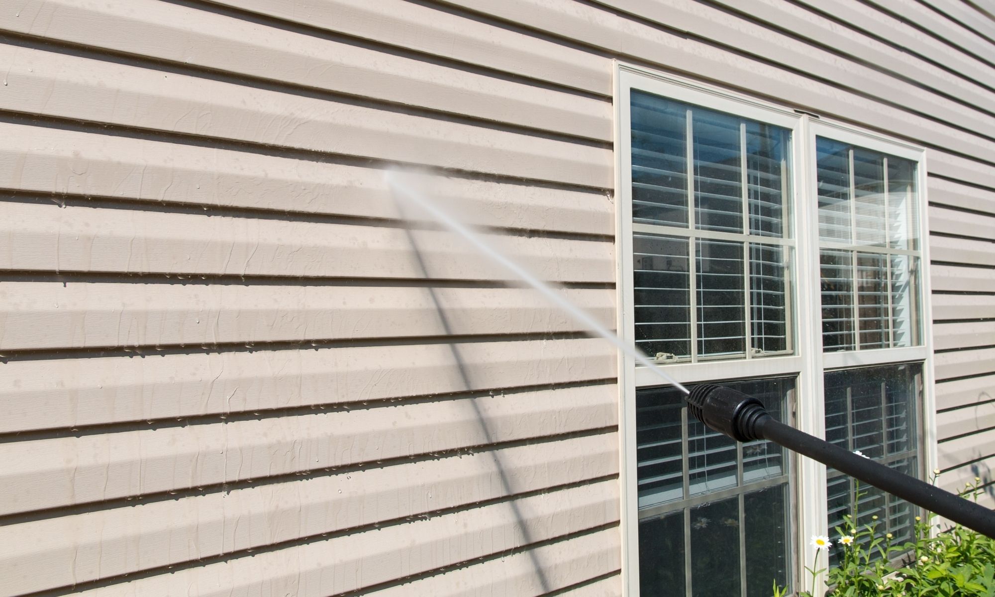 A person uses a pressure washer for house washing, cleaning beige siding near a window with white shutters.