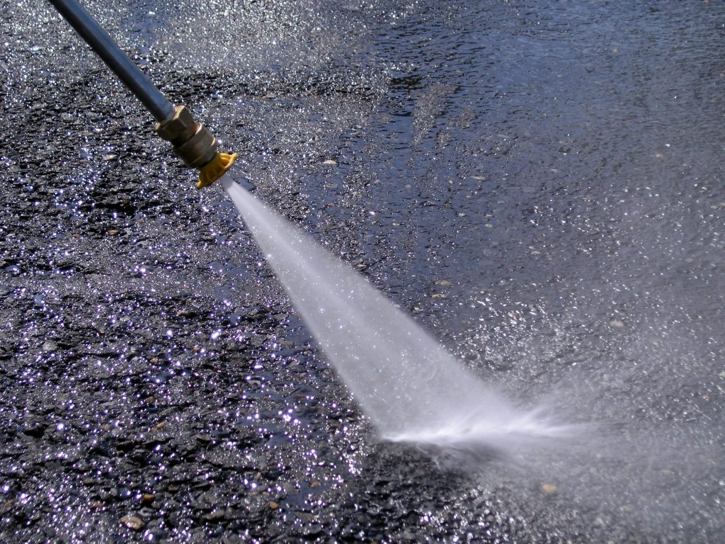 Pressure washer nozzle sprays strong water stream onto wet, textured asphalt, perfect for pressure washing surfaces.