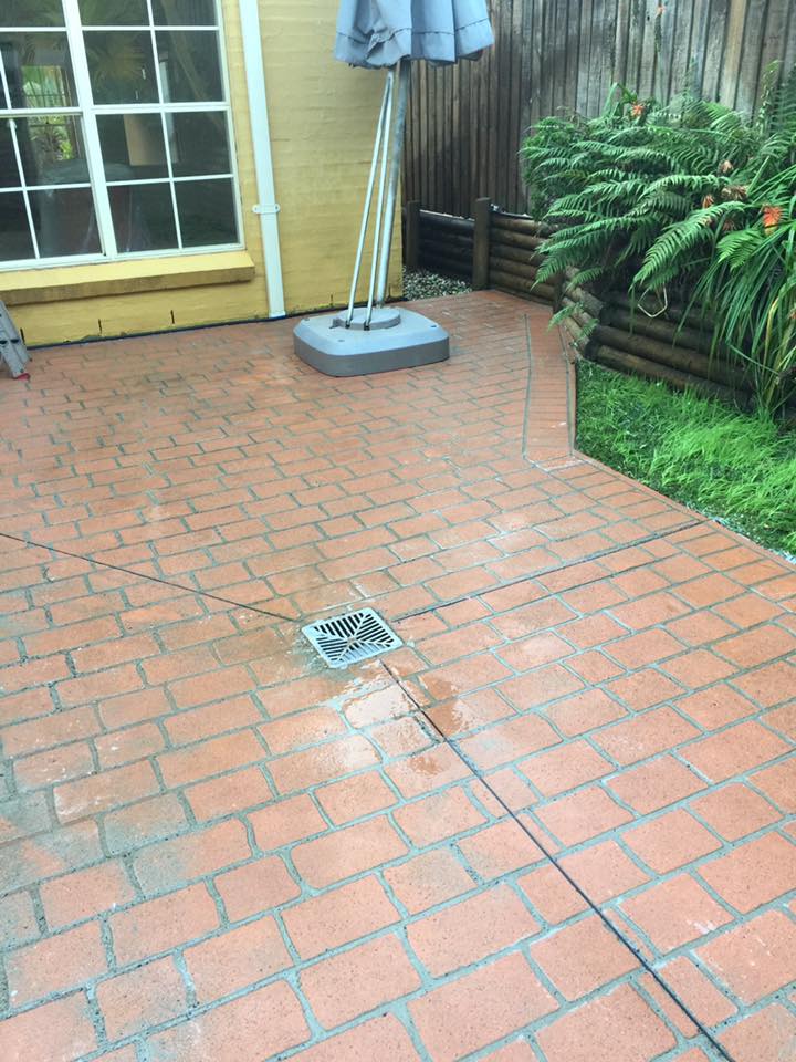 Red brick patio with central drain, pressure-washed wet patches, umbrella on stand, and plants along wood fence.