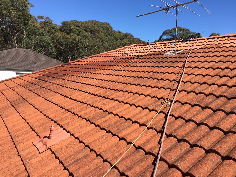 Red tiled roof with TV antenna and safety rope, ideal for gutter cleaning; trees and blue sky in the background.