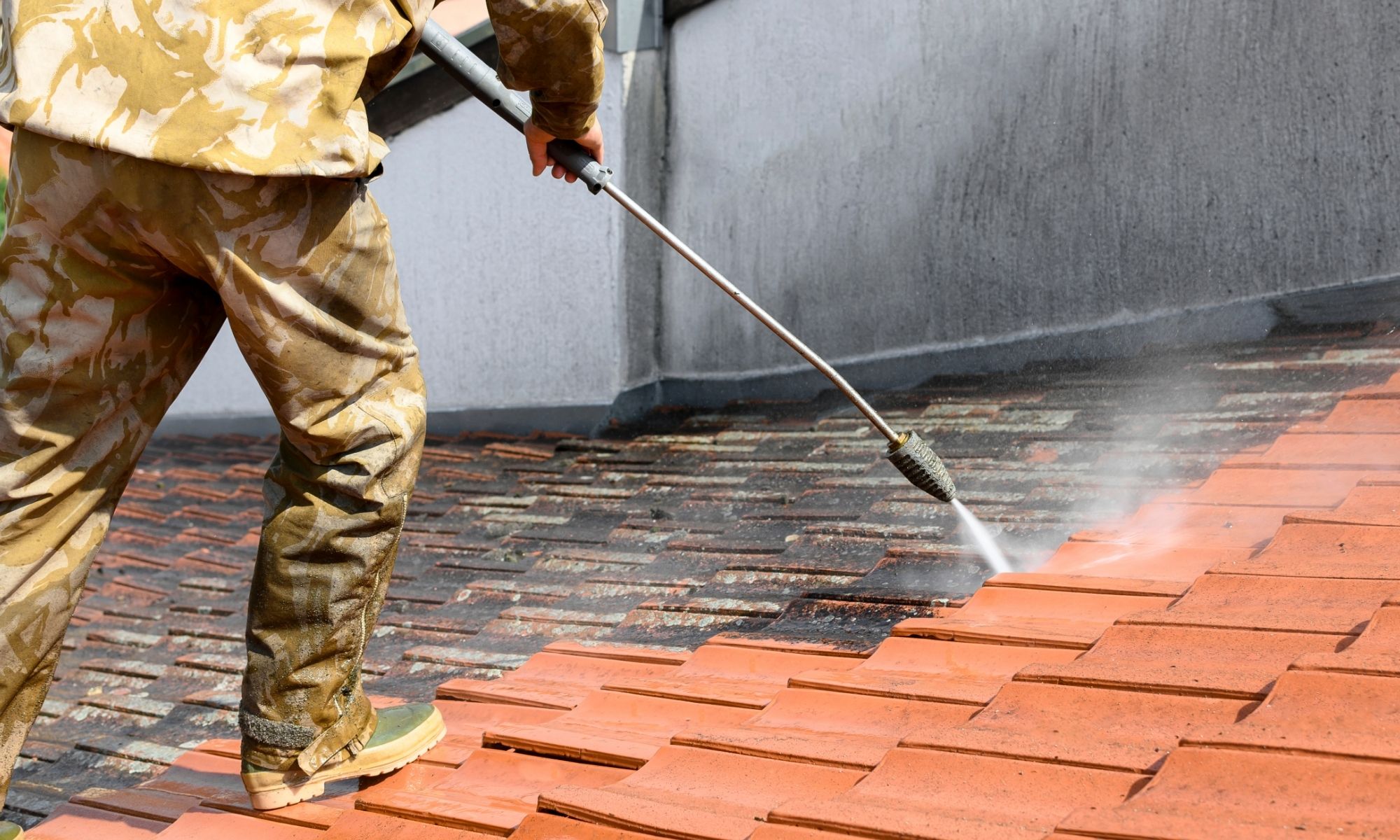 Person in camouflage clothing pressure washes dirt and moss off a tiled roof, revealing clean red tiles.