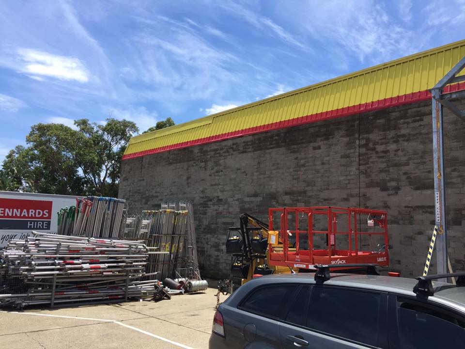 Outdoor scene with stacked scaffolding equipment, red and yellow building, parked car, and Sénnards Hire sign.