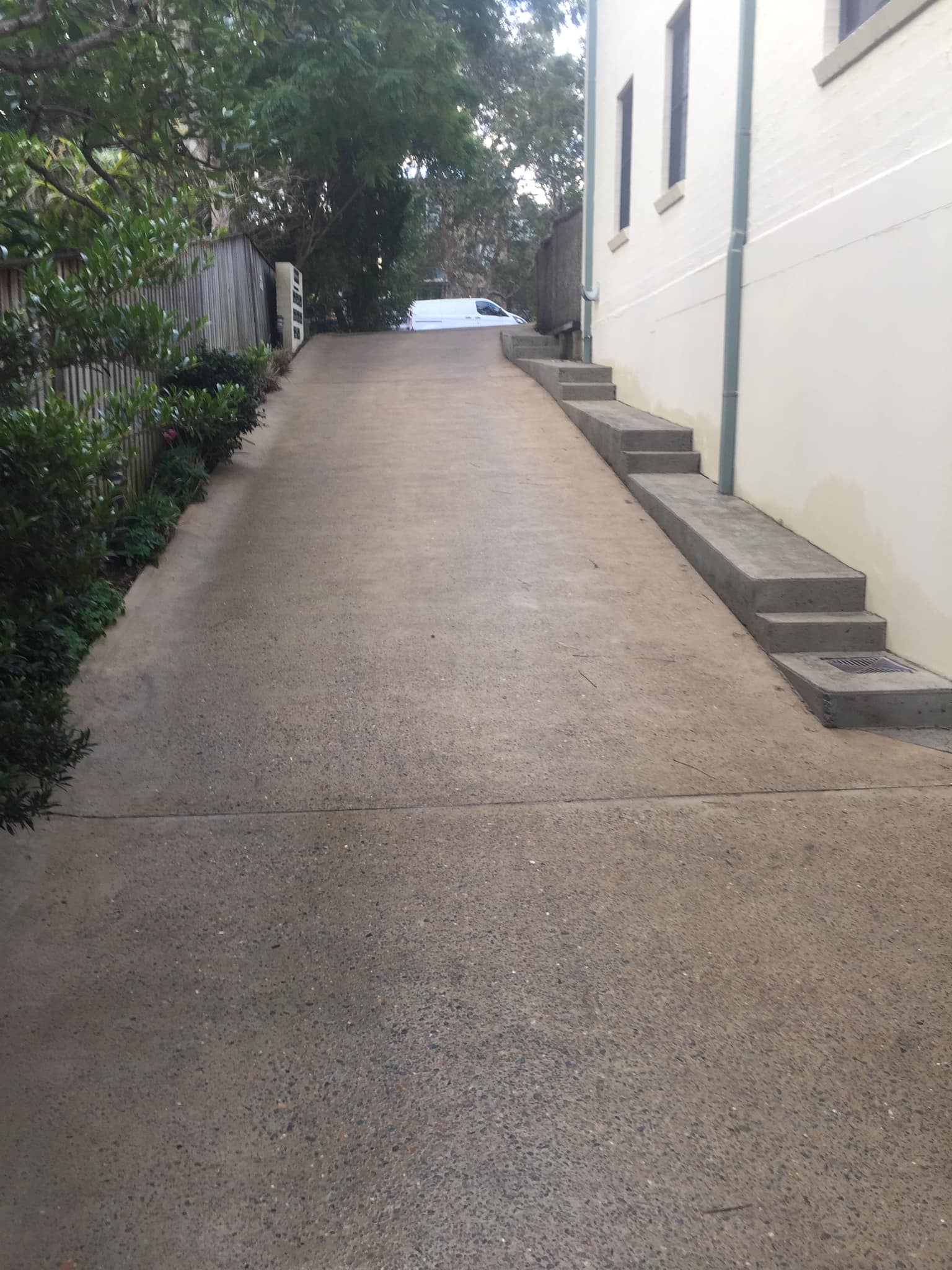 Sloped concrete driveway ideal for pressure cleaning beside a white building, greenery left, white car at top.