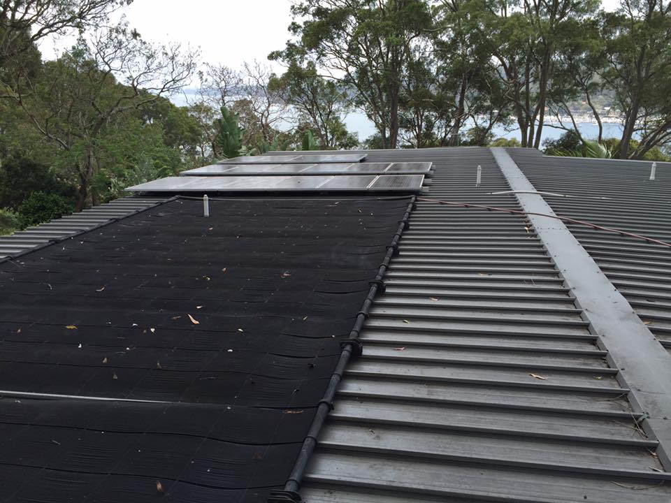 Solar pool heating mats and panels on a metal roof near trees, stressing the need for gutter cleaning; water in background.