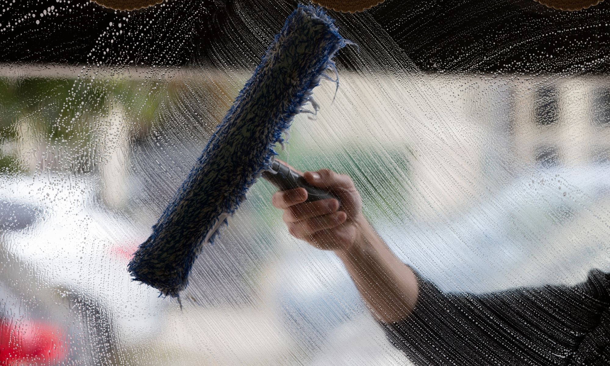 Window cleaning with a blue squeegee on glass, soap streaks visible, blurred cars and buildings in the background.