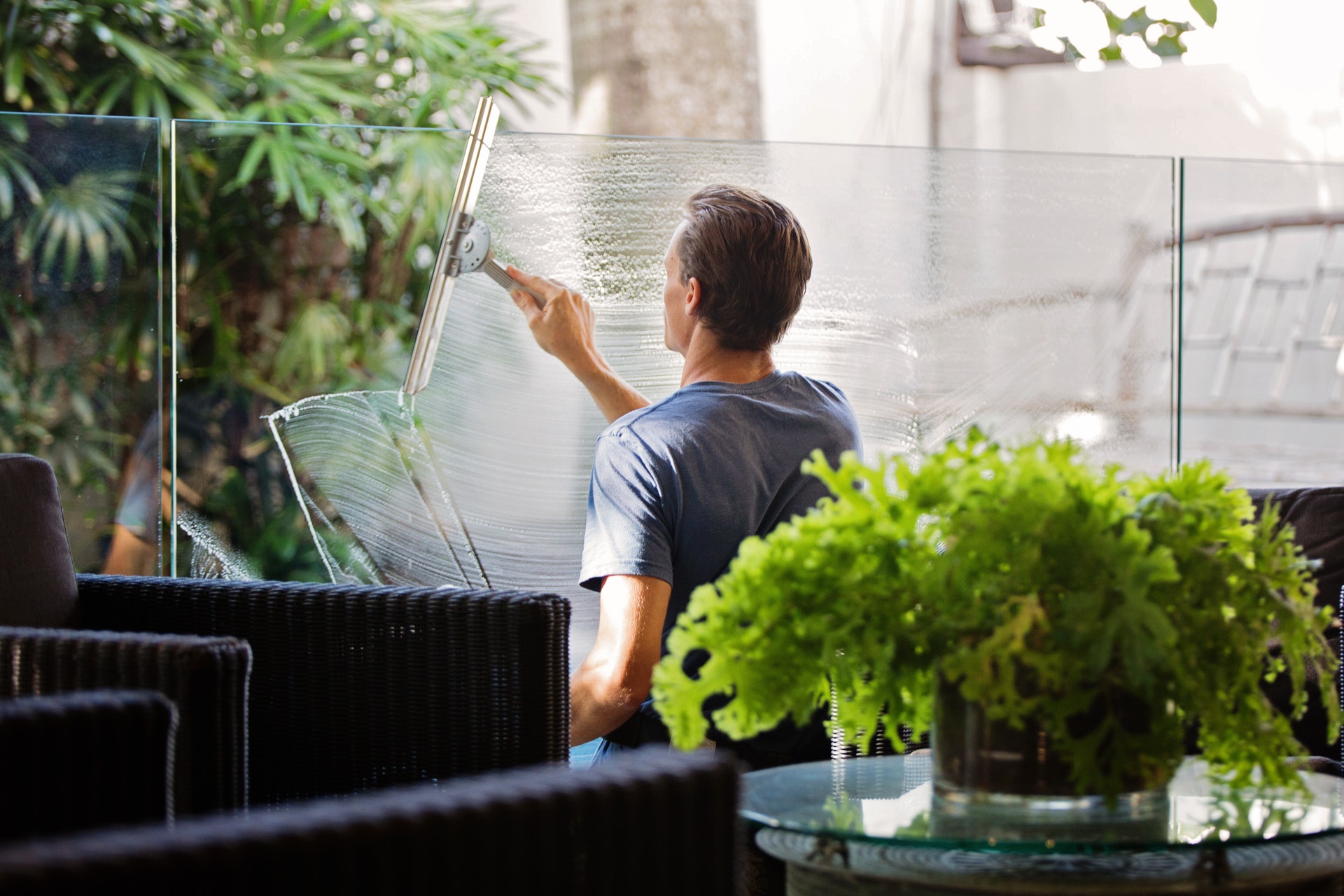 Window cleaning with a squeegee on a large glass window, outdoor wicker furniture and lush green plants nearby.