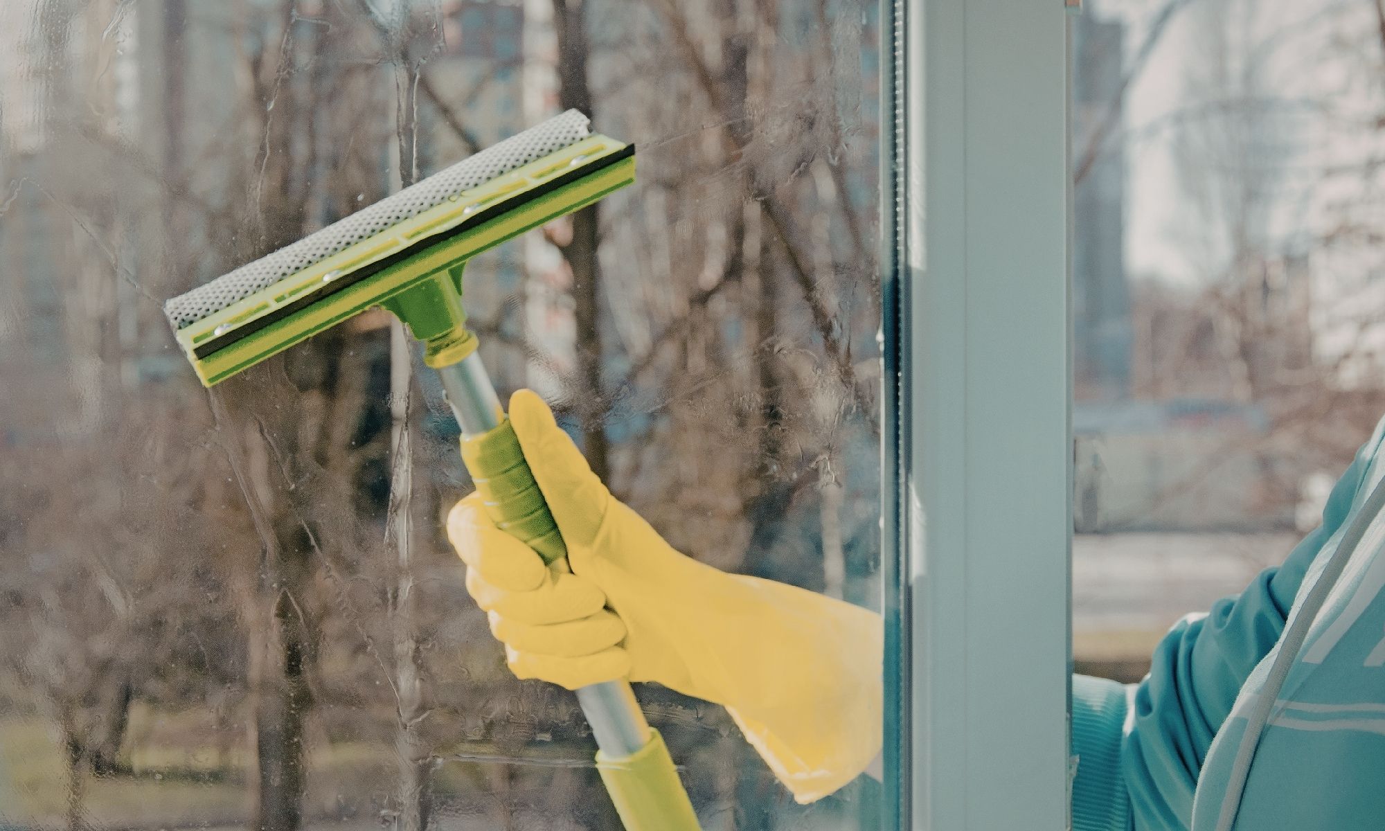 Person wearing yellow gloves using a green squeegee to clean a glass window