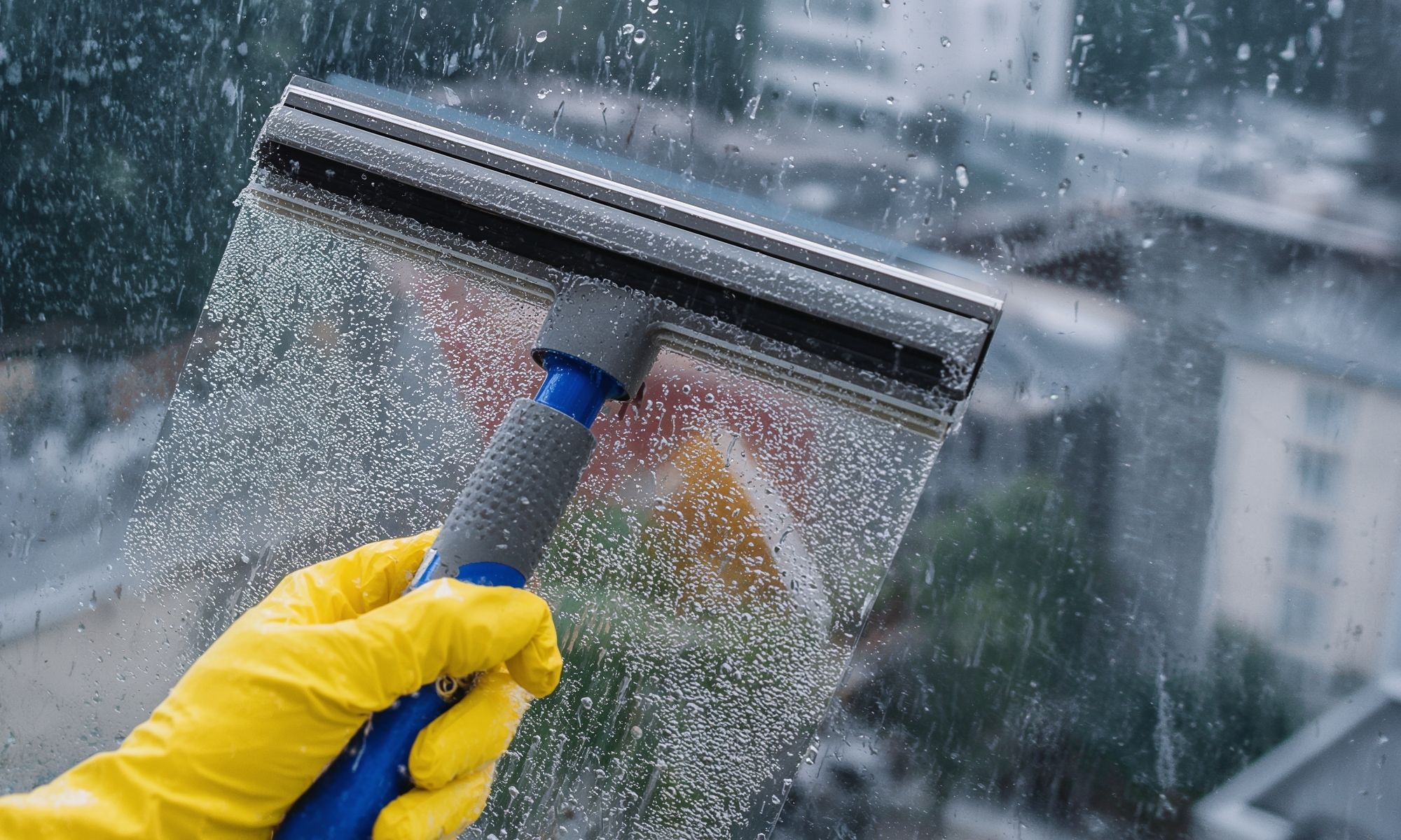Person wearing yellow gloves grips a blue window squeegee while rain beads on the glass