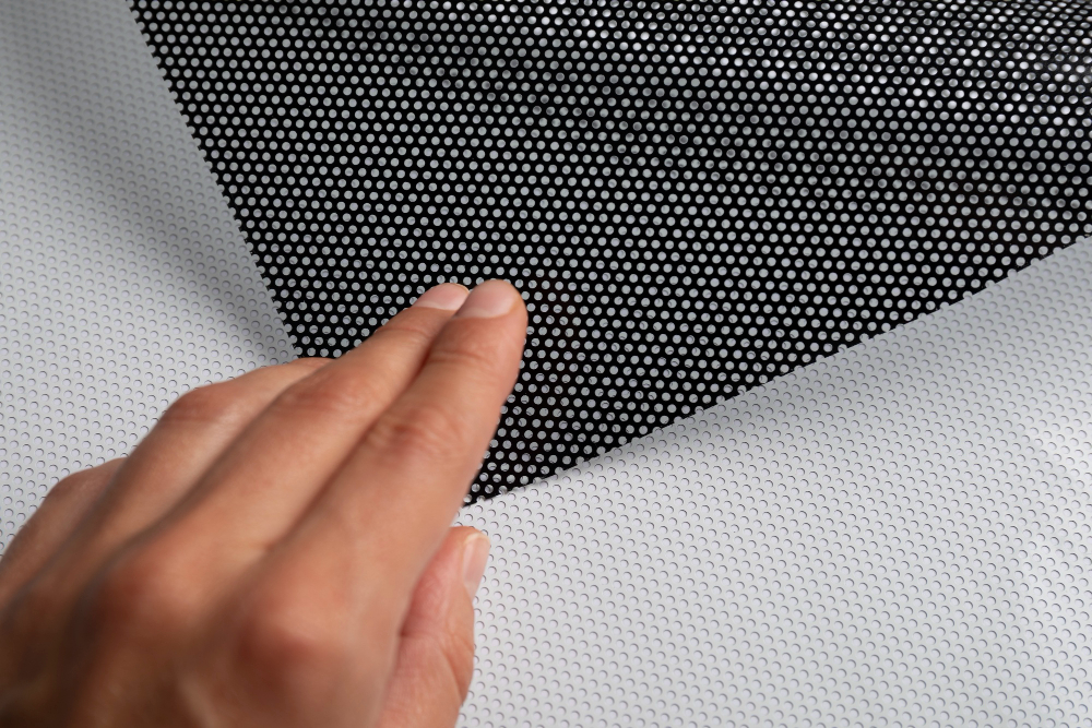 A hand lifts black perforated material, like a Window Fly screen, revealing similar white perforated surface below.